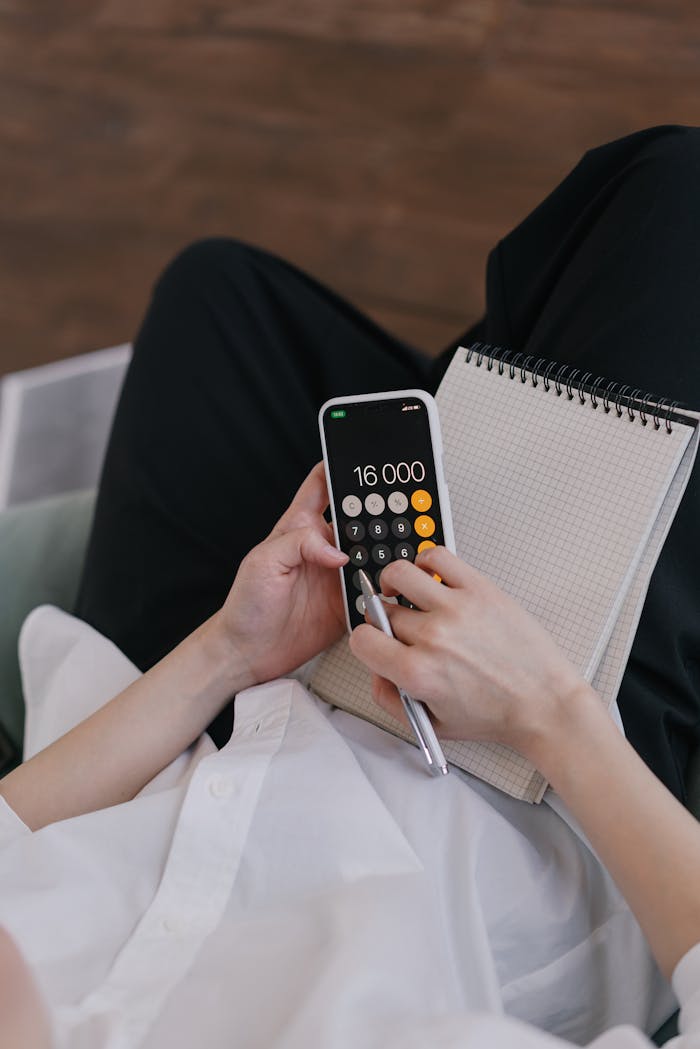 Close-up of a person using a smartphone calculator while planning finances with a notebook.